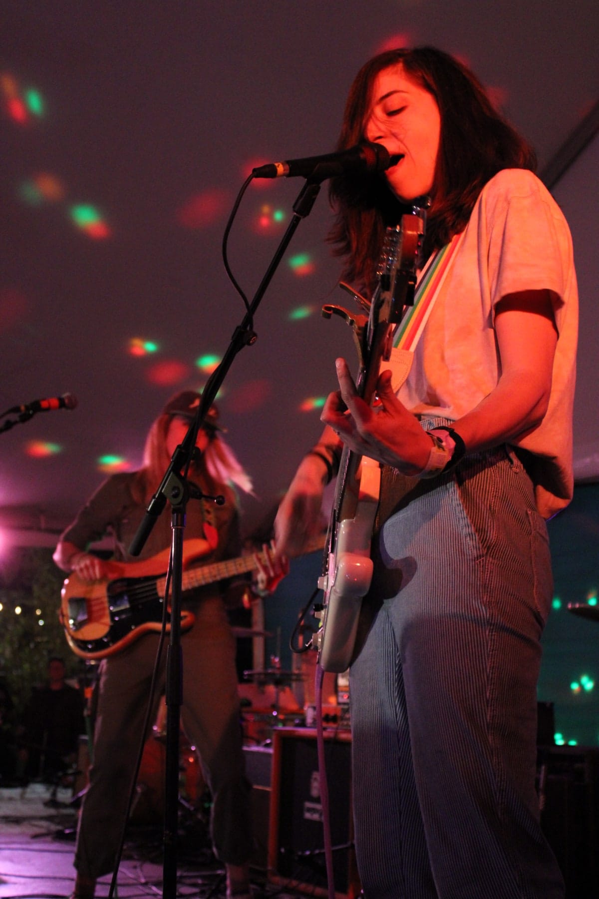 A guitarist sings into a microphone in warm stage light, with another musician behind.
