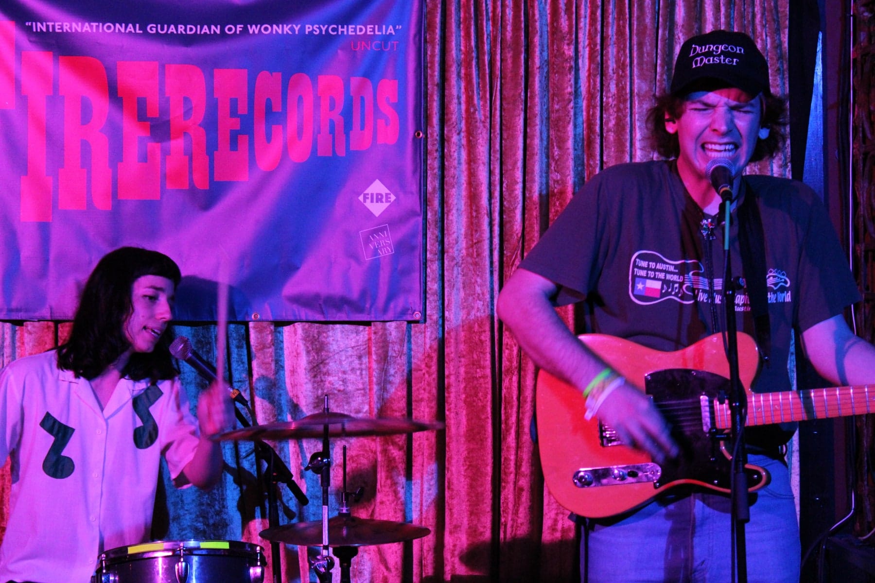 Two musicians perform side by side under pink and purple stage light.