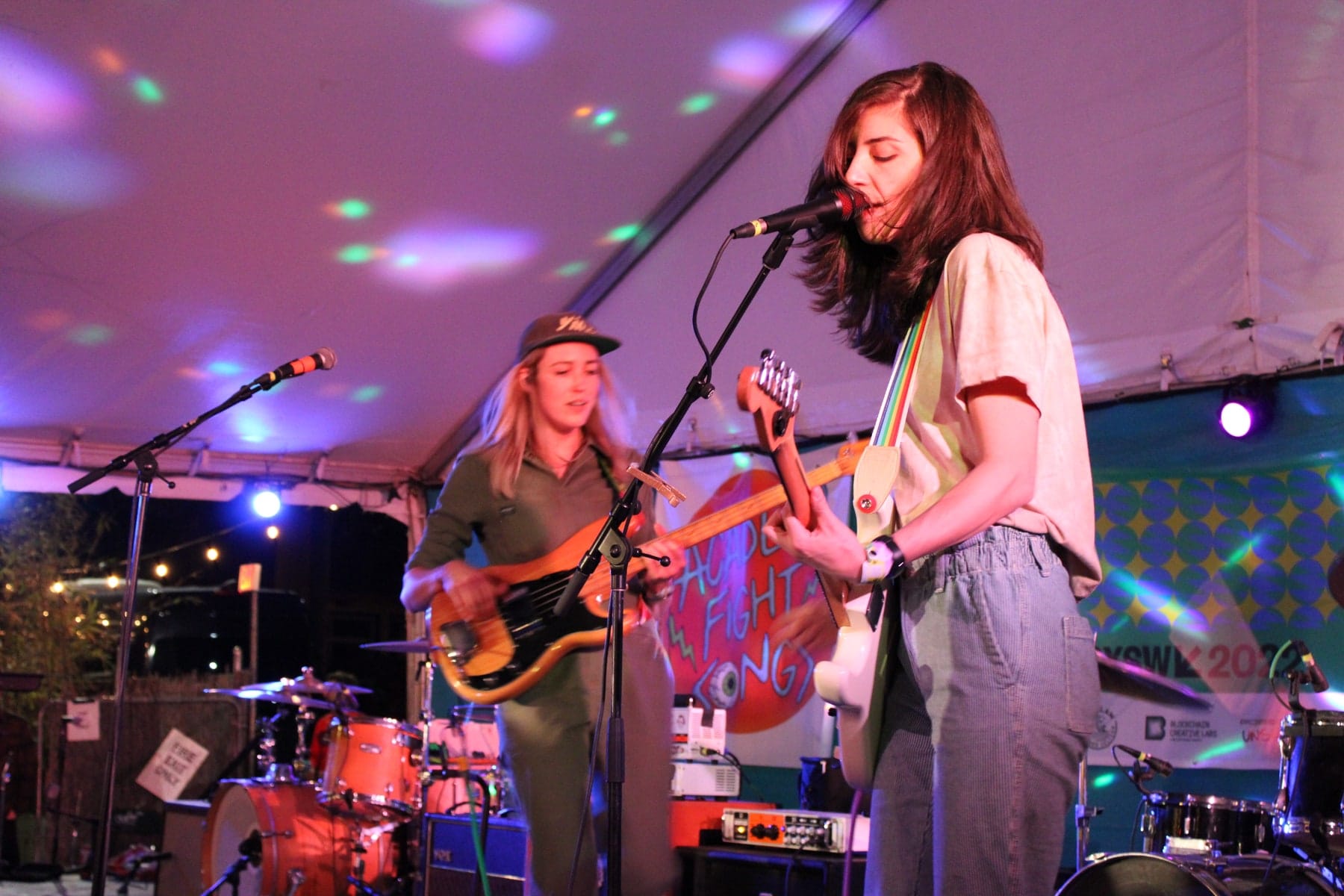 A wider view of a singer and guitarist, bassist and drummer under colored light.