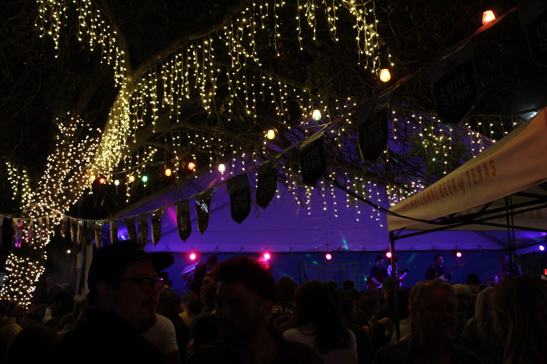 Audience silhouettes and a lit stage beneath strings of lights at night.